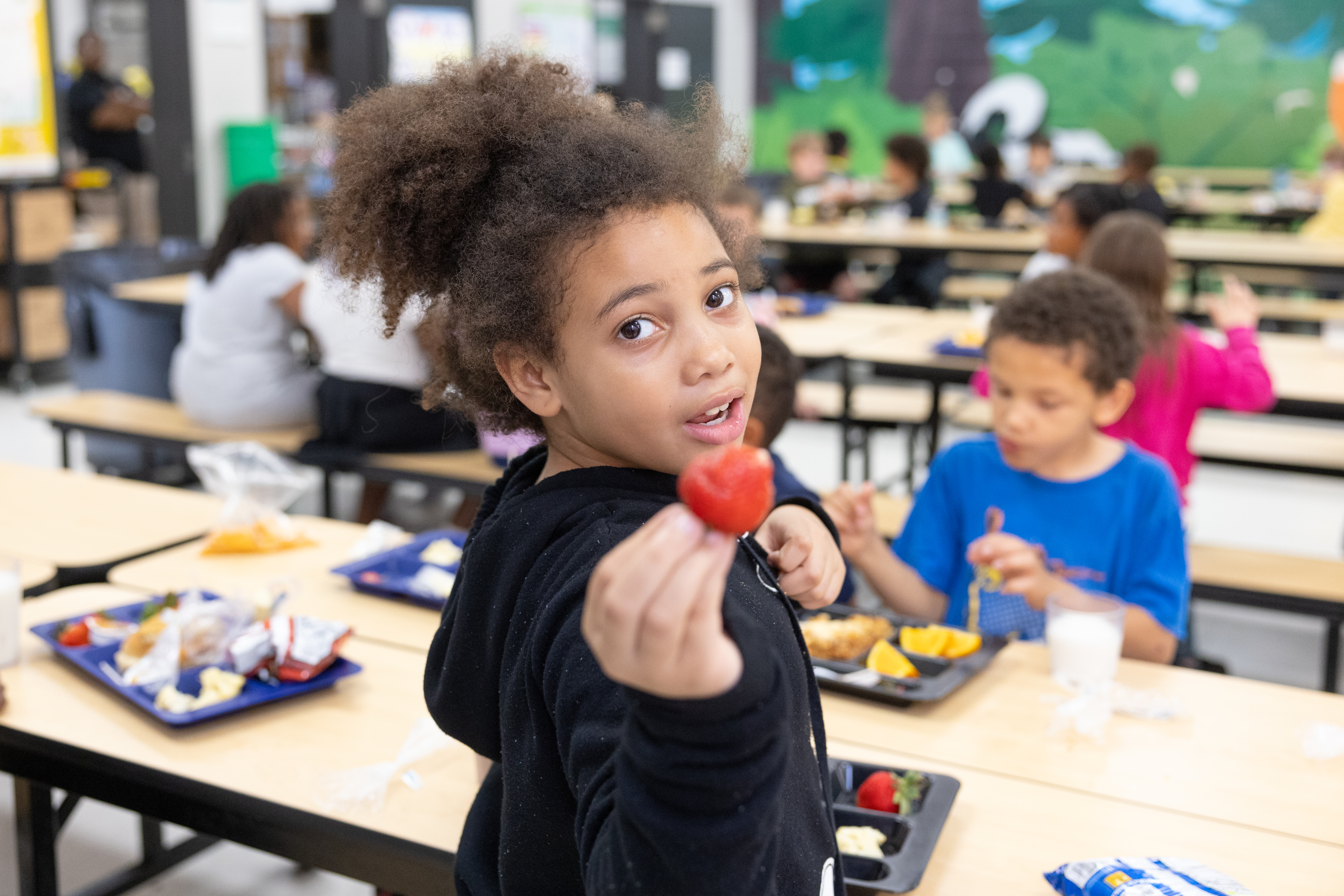 A young black girl sits at a lunch table turned around and shows a strawberry to the camera.