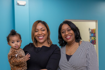 Two black women stand in the center of the image smiling. The woman on the left is holding a smiling young child. 