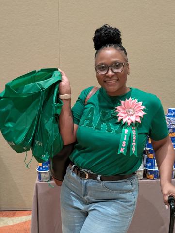 woman dressed in green AKA shirt and pink rosette holds up green grocery bags packed for families