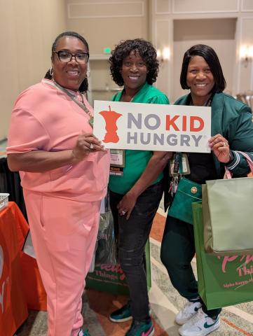 three women dressed in pink and green hold up a No Kid Hungry sign